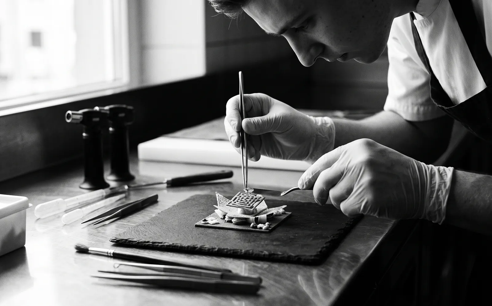 A gloved chef using tweezers and precision tools to assemble an intricate technical dish