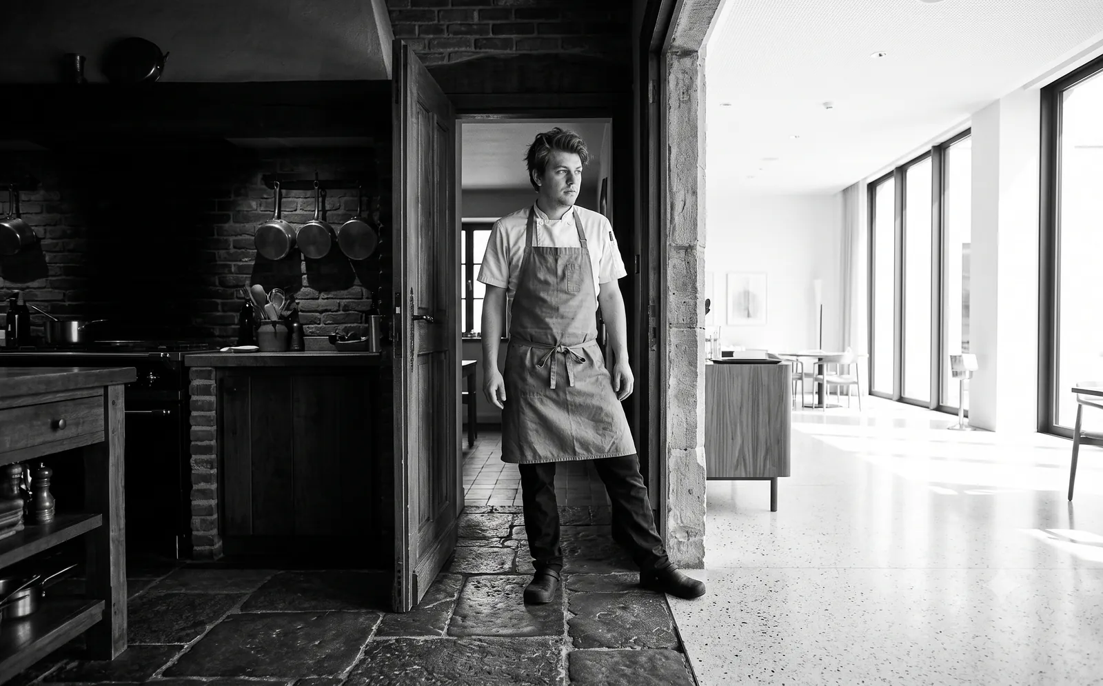 A chef pausing in a doorway between an old stone kitchen and a bright modern dining room
